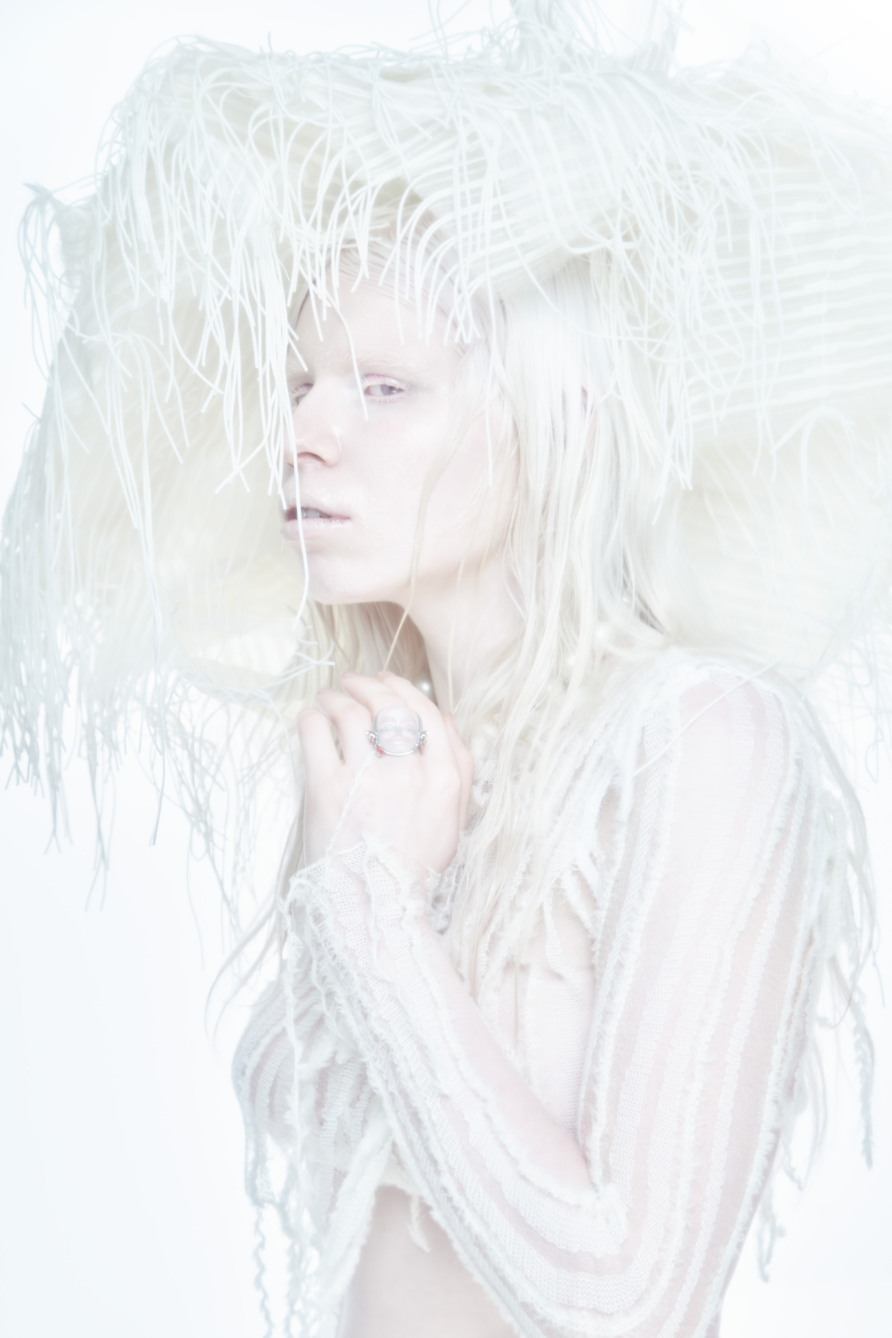 Monochrome portrait of a woman wearing an elaborate feathered hat and white textured sheer top against a white background.