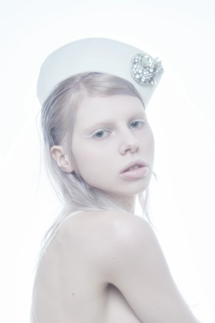 Model wearing a white pillbox hat adorned with crystal brooch against a bright, high-key white seamless backdrop for Flanelle Magazine.