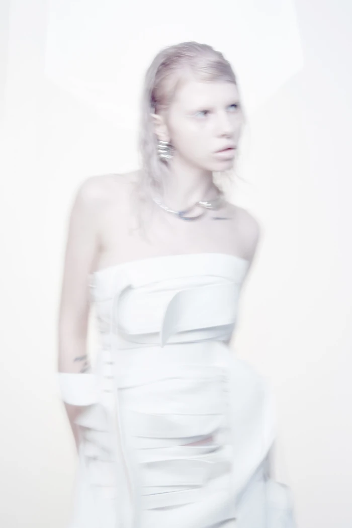 Model with blonde hair and pale styling wearing a structured white strapless dress against a bright white background.