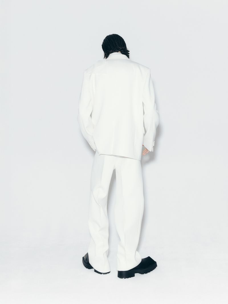 Back view of an adult male model in white suit and black shoes against a white background in a fashion studio setting.