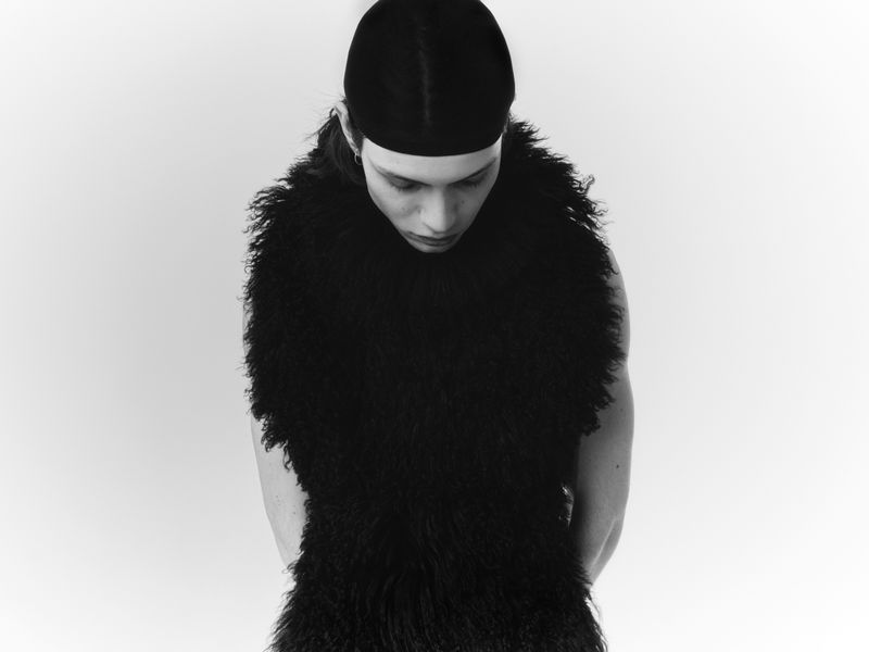 Monochrome portrait of person wearing a cap and textured sleeveless garment, looking down against a bright white backdrop.