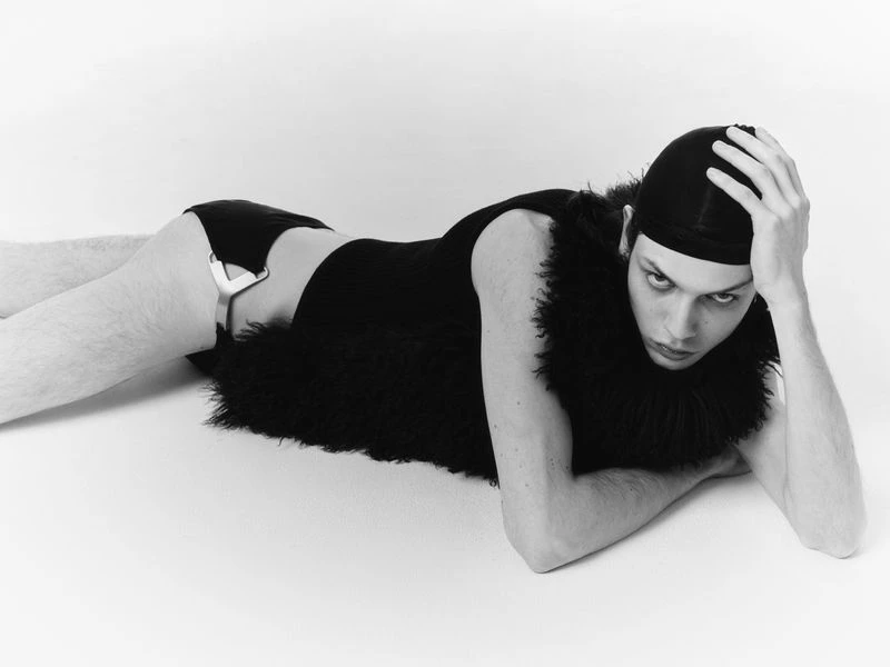 Fashionable male model lying on a white surface wearing a black vest, briefs, and a head covering poses with hand on his head.