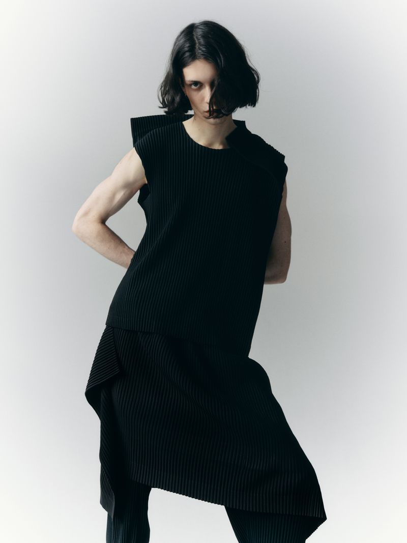 Model poses in a pleated black sleeveless tunic and pants against a plain white backdrop in a fashion editorial style shot.