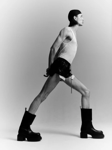 Fashionable, athletic-build adult male in a sleeveless shirt, shorts, and boots stretches in a photo studio with soft, even lighting.
