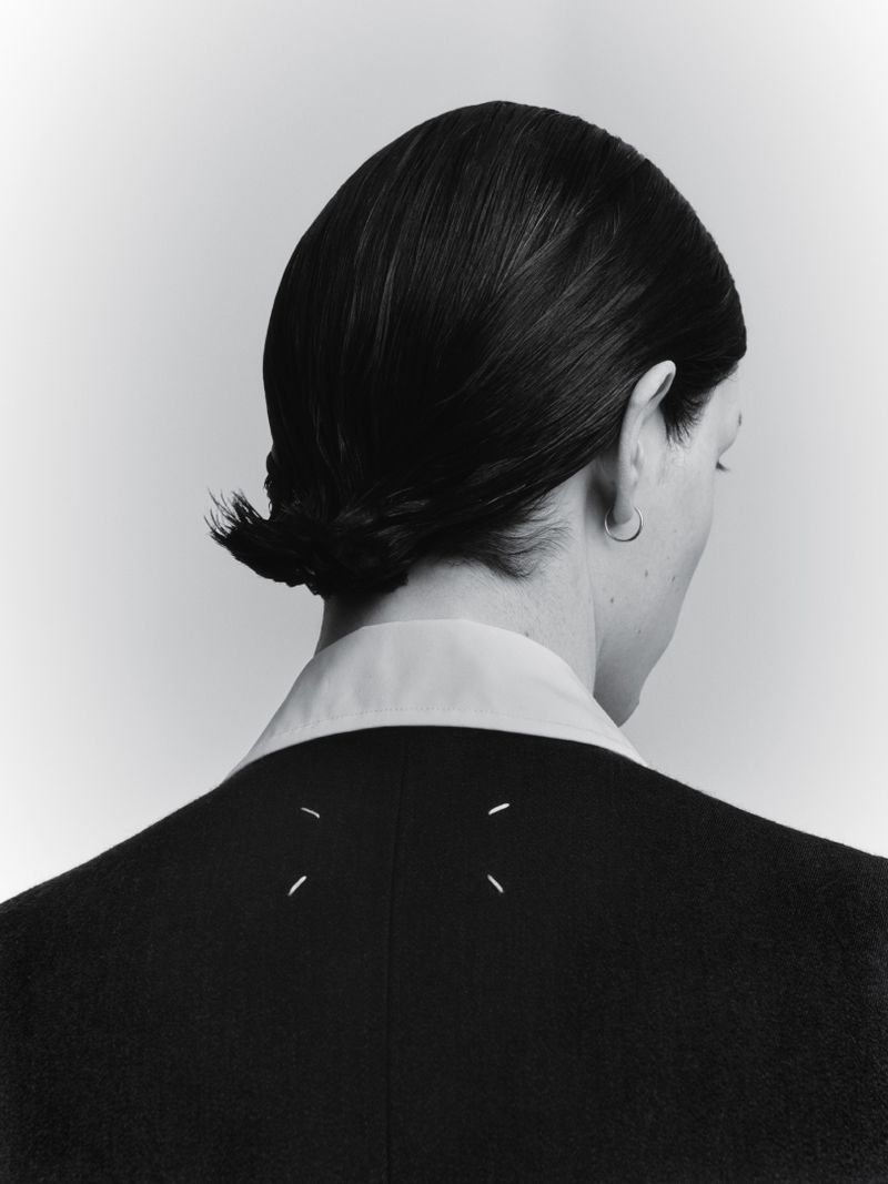 Fashion shot: Person with dark hair in bun, wearing a dark blazer with white stitches on the back, and a white collared shirt.