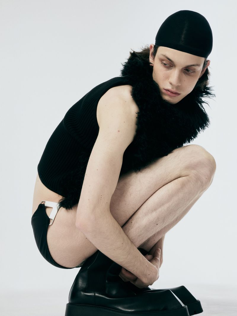 Studio shot of a crouched male model wearing a black beanie, sleeveless top, and platform boots in a fashion-forward, neutral setting.