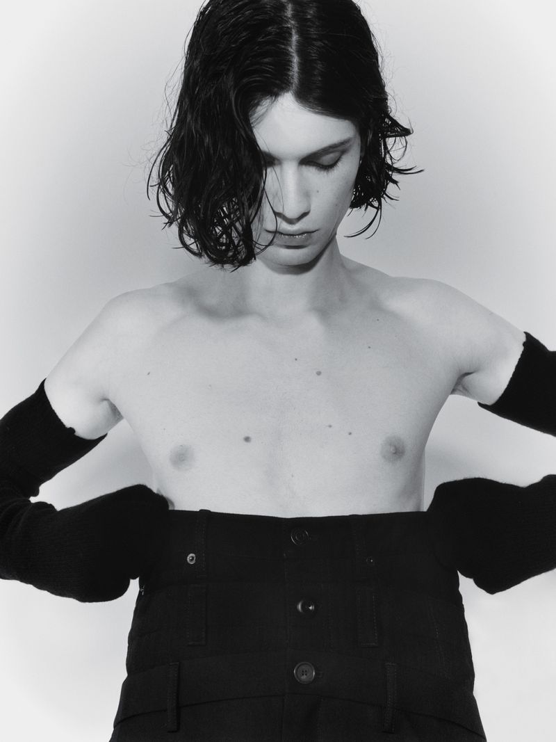 Monochrome portrait of a shirtless person with dark hair, wearing black pants and arm sleeves, looking down in a studio setting.