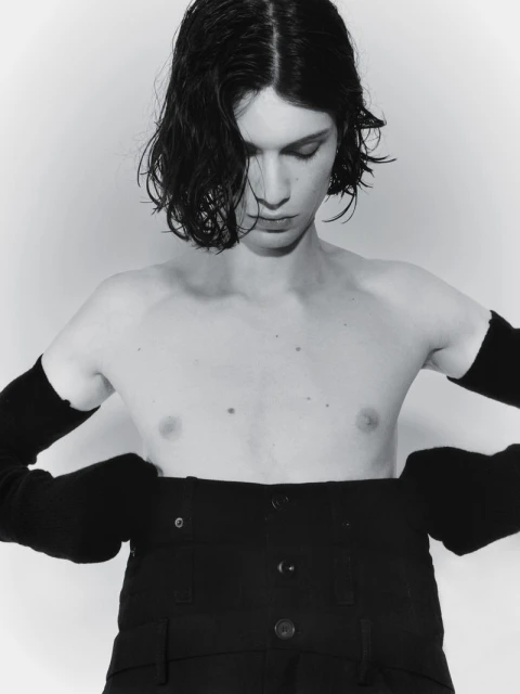 Monochrome portrait of a shirtless person with dark hair, wearing black pants and arm sleeves, looking down in a studio setting.