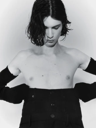 Monochrome portrait of a shirtless person with dark hair, wearing black pants and arm sleeves, looking down in a studio setting.