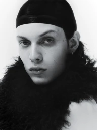 Close-up monochrome portrait of a model wearing a dark fitted cap and textured fuzzy collar against a white background.