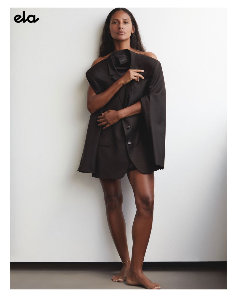 Brunette adult woman poses indoors with bare shoulders, wearing an oversized dark blazer, against a white wall in a fashion editorial.