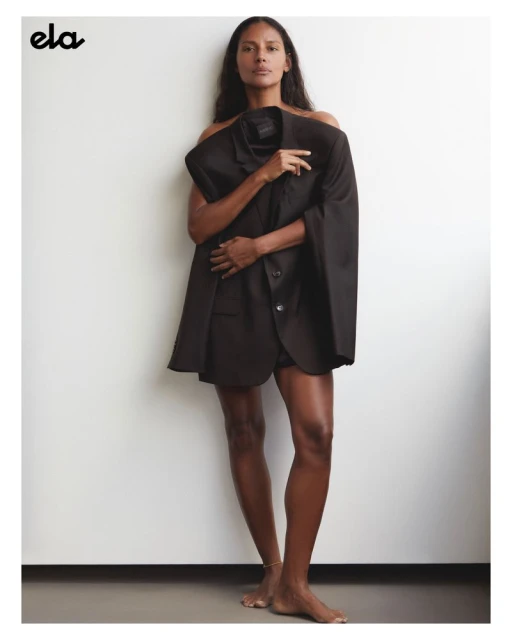 Brunette adult woman poses indoors with bare shoulders, wearing an oversized dark blazer, against a white wall in a fashion editorial.