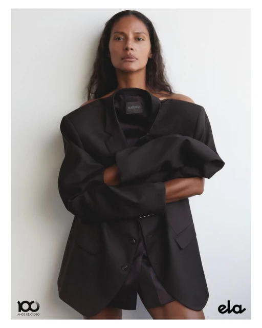 A dark-haired woman poses in front of a neutral background, partially covered by an oversized black blazer, for a fashion editorial.
