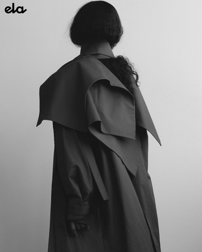 Monochrome fashion shot from behind of an adult female with dark hair wearing a dark, oversized trench coat against a light backdrop.