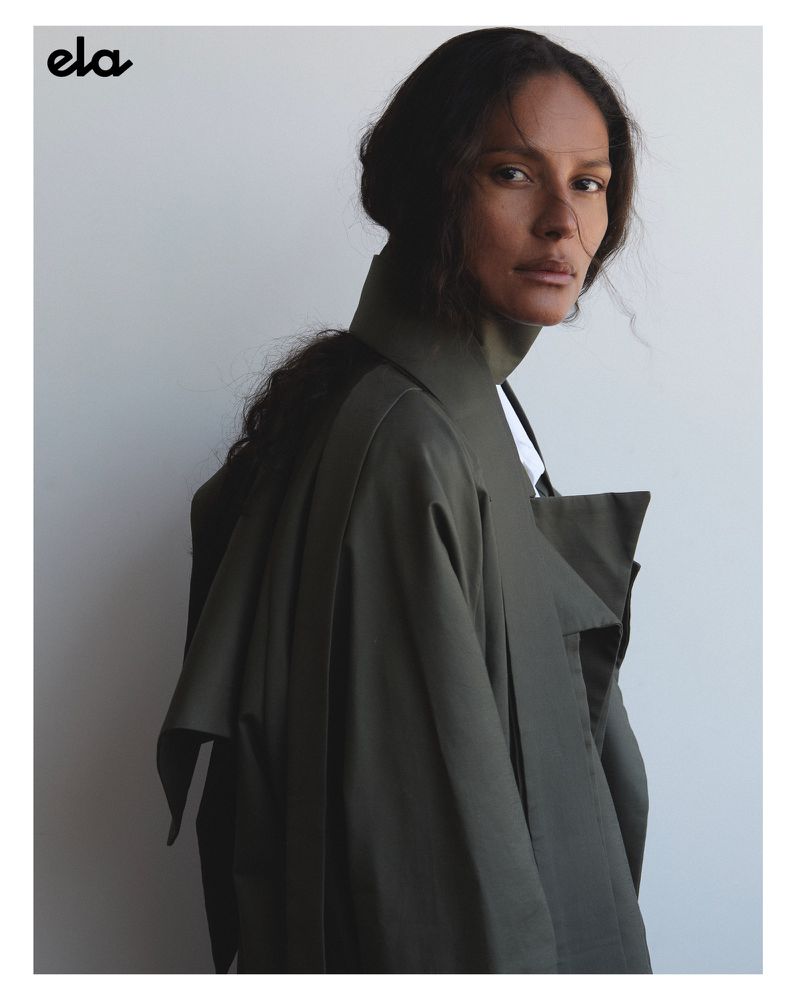 Adult female model in olive trench coat poses in a studio setting. Neutral background with natural light highlighting features.