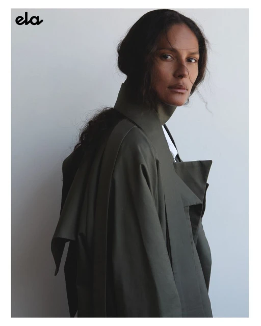 Adult female model in olive trench coat poses in a studio setting. Neutral background with natural light highlighting features.