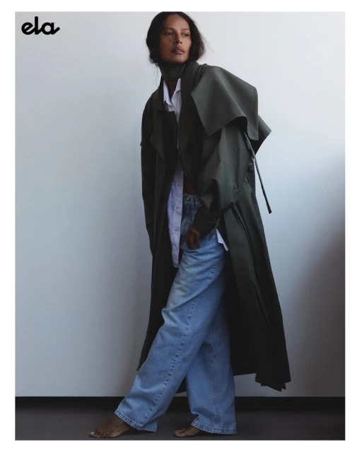 A fashion shot shows a biracial adult female in a green coat, blue jeans, and no shoes, posing in front of a white wall in a studio.