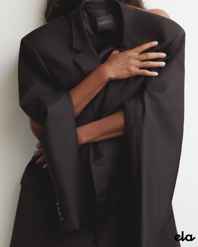 A woman embraces an oversized black blazer, creating a chic fashion statement against a neutral backdrop with an 'ela' logo.