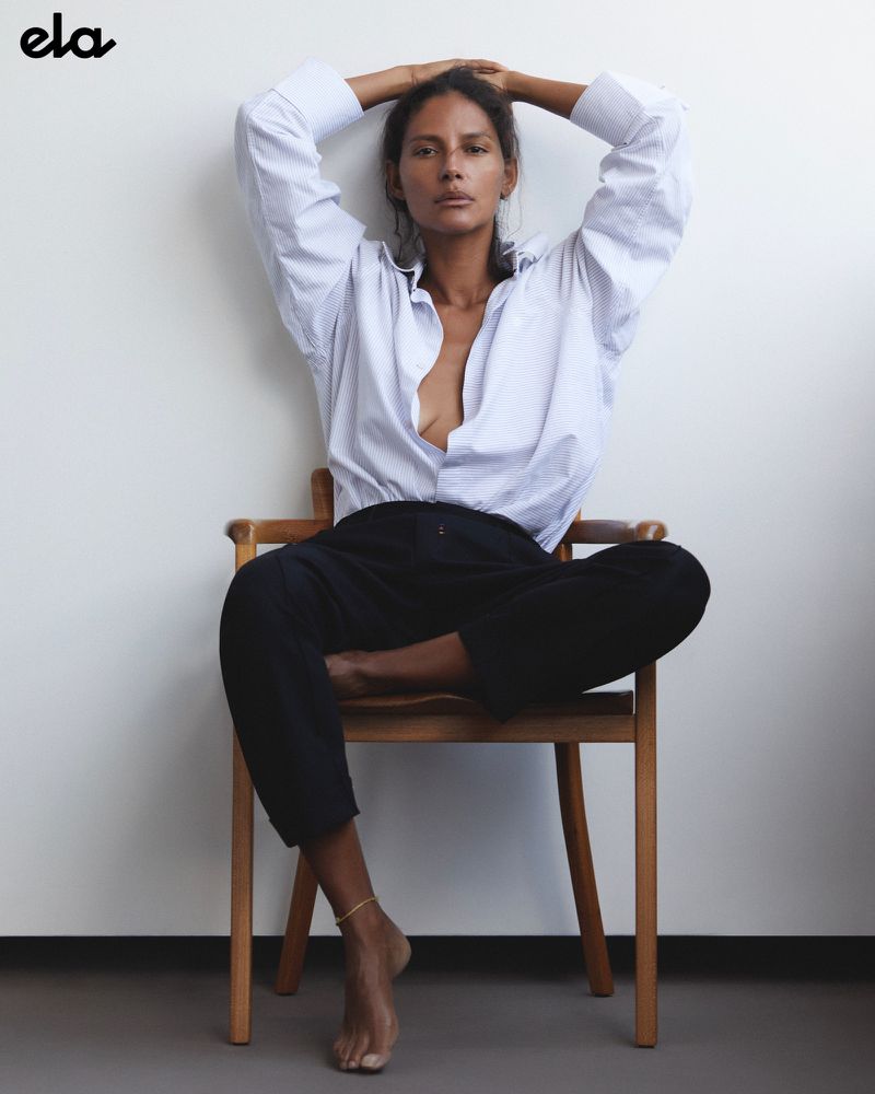 Brunette woman in an unbuttoned blue-striped shirt and black pants sits cross-legged on a wooden chair against a white wall.
