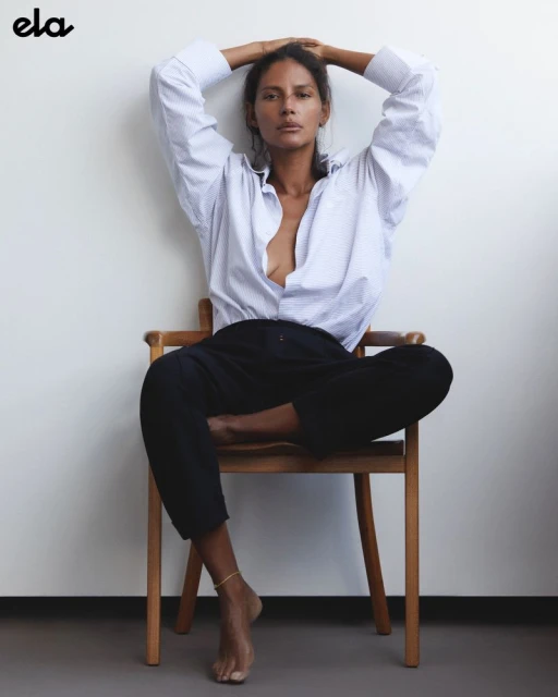 Brunette woman in an unbuttoned blue-striped shirt and black pants sits cross-legged on a wooden chair against a white wall.