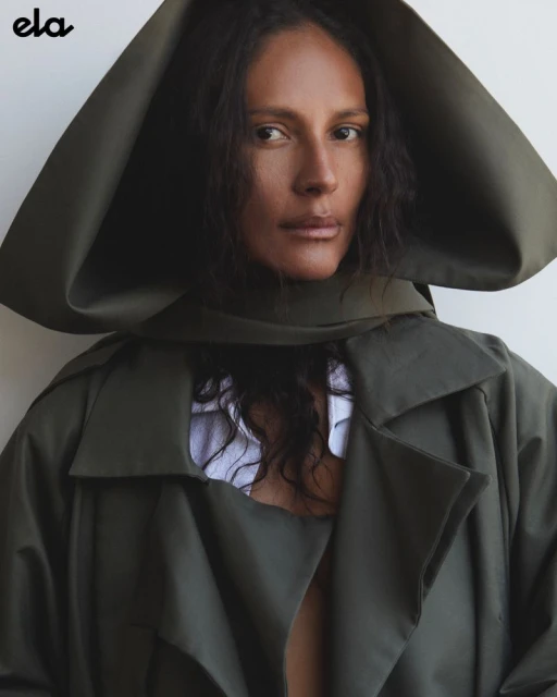 Portrait of adult female model wearing olive green trench coat with oversized hood against a neutral backdrop in a studio.