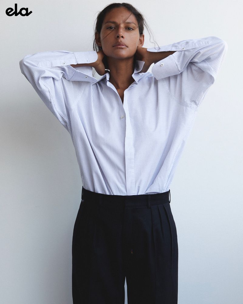 An adult woman poses indoors wearing a blue and white striped button-down shirt and black trousers against a neutral wall.