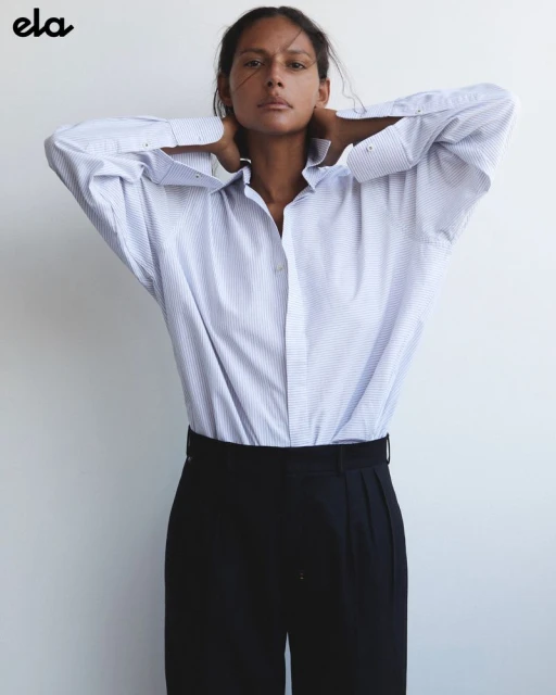 An adult woman poses indoors wearing a blue and white striped button-down shirt and black trousers against a neutral wall.