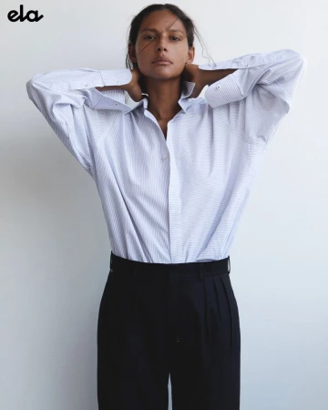 An adult woman poses indoors wearing a blue and white striped button-down shirt and black trousers against a neutral wall.
