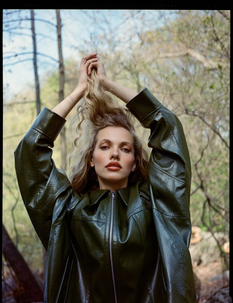 A young woman with blonde, curly hair raises her hands above her head, posing in a green, shiny jacket against a wooded background.