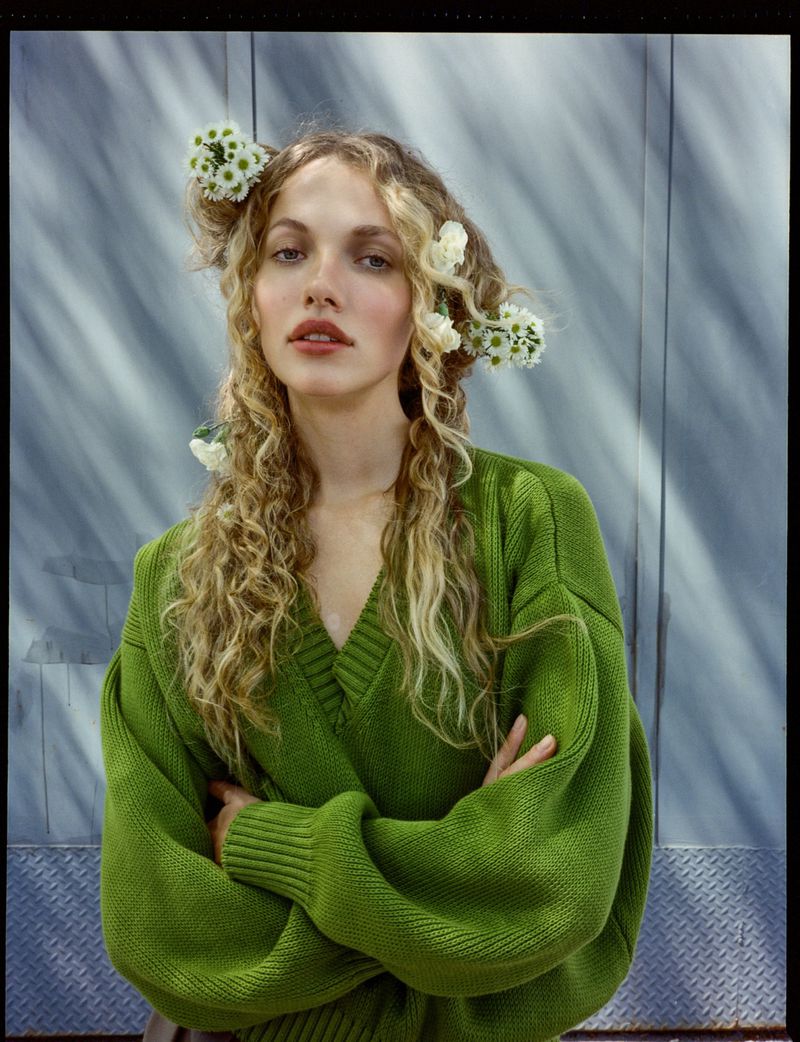 Portrait of a woman with curly blonde hair adorned with white flowers, wearing a green v-neck sweater and posing indoors.