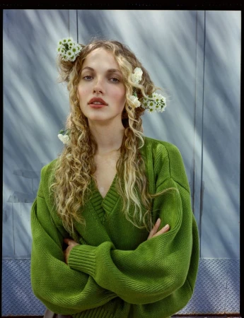 Portrait of a woman with curly blonde hair adorned with white flowers, wearing a green v-neck sweater and posing indoors.