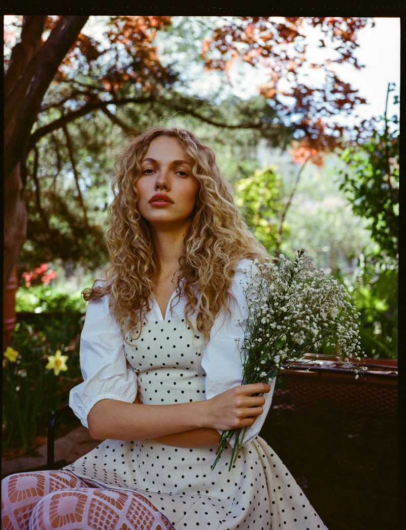 Blonde woman in polka-dot dress poses in a garden with baby's breath flowers, styled with patterned tights and soft, natural light.
