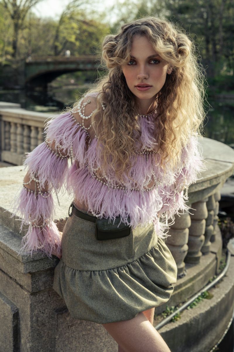 Young woman in an outdoor fashion shot, wearing a lilac feathered top and an olive green skirt, posing near a stone railing and a bridge.