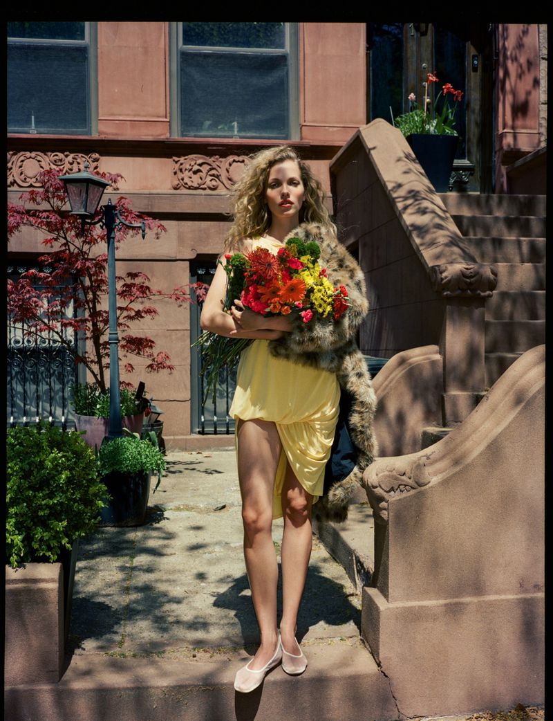 Woman in yellow dress with flowers and fur stole standing on steps of brownstone building in urban setting on sunny day.