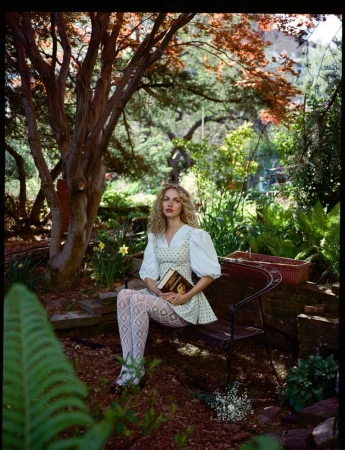 A young woman with curly blonde hair is sitting on a bench in a garden setting, holding a book and wearing a patterned dress and tights.