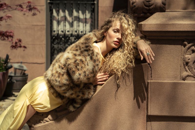 Elegant woman with blonde curly hair, wearing a spotted fur coat and yellow dress, posing on brown stone steps, in outdoor setting.