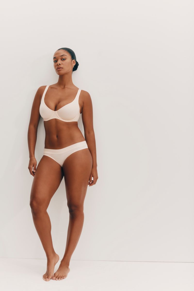 Woman in matching beige bra and underwear set poses against a clean white background in a studio shot, looking at the camera.