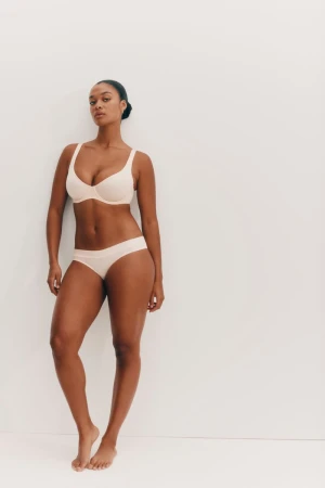 Woman in matching beige bra and underwear set poses against a clean white background in a studio shot, looking at the camera.
