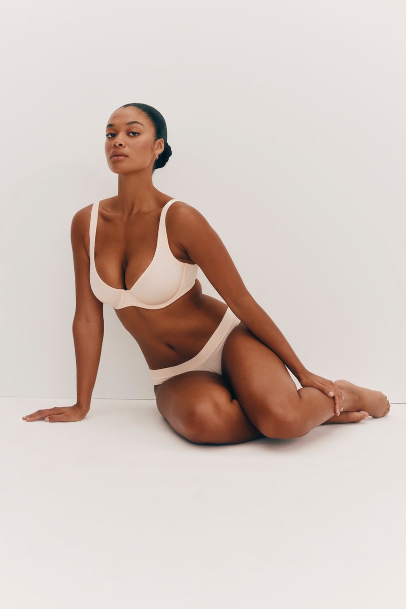 A slender adult woman poses indoors wearing a matching light pink bra and underwear set against a bright white background.