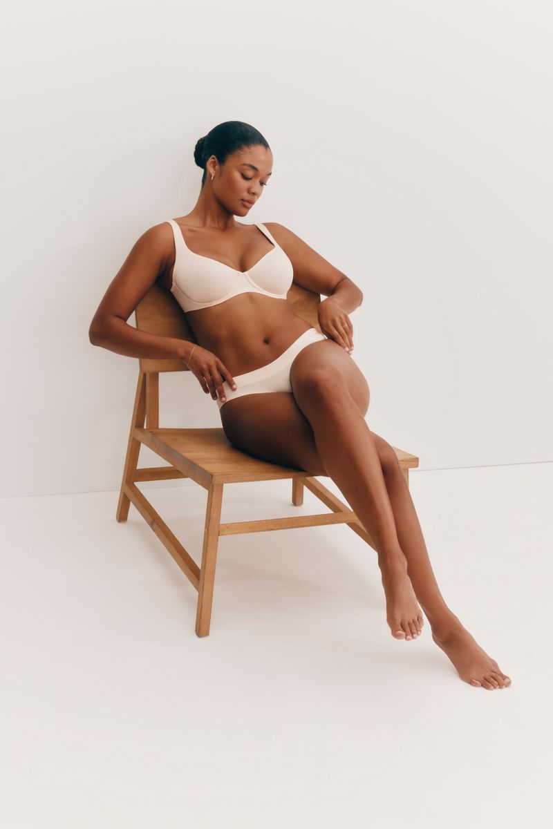 Woman in lingerie seated on a wooden chair against a white background in a medium shot.