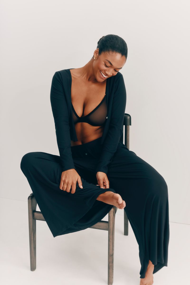 Smiling woman in black lounge wear set sitting on chair in studio, fashion pose, indoor setting, lit background. Beauty shot.