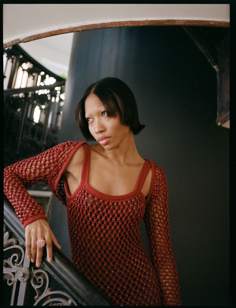 Indoor portrait of a Black woman in a red knit dress, standing on a dark staircase with light stone and decorative metalwork.