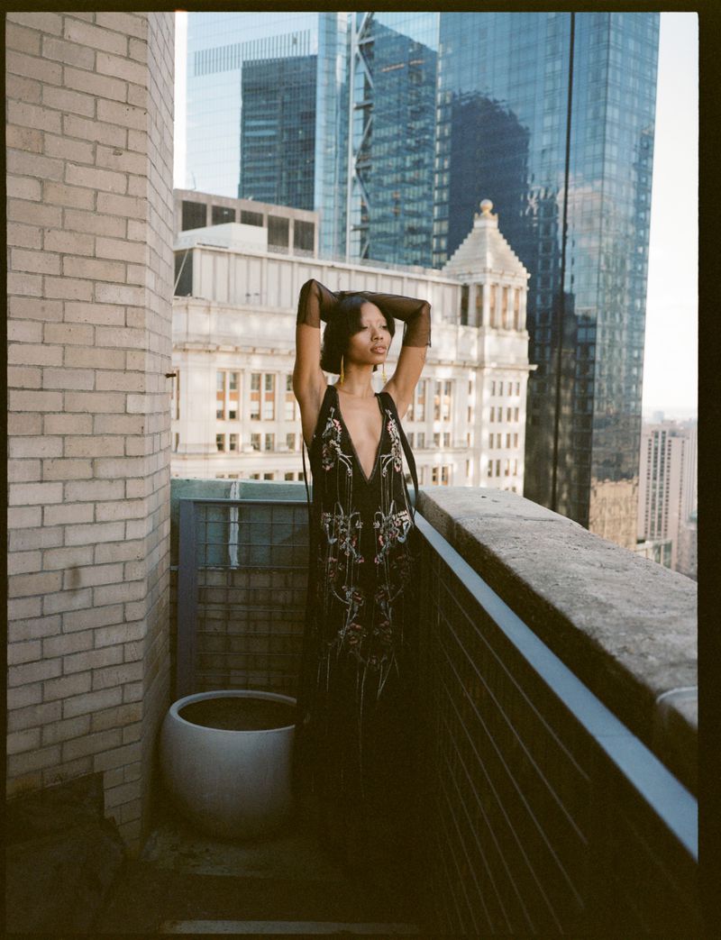 Woman in a dark dress posing on a rooftop with skyscrapers in the background, wearing sheer gloves and standing next to a planter.