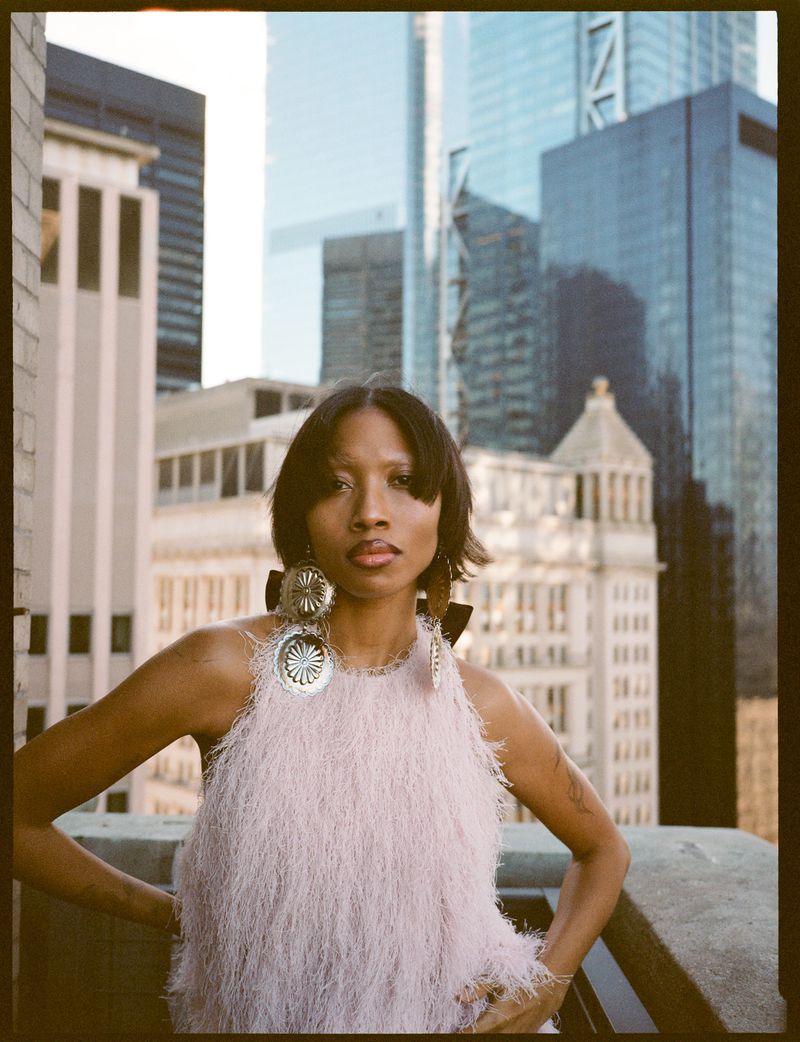 Adult woman with short brown hair poses in a pink, fringed dress, large earrings, and a city skyline backdrop. Beauty and Fashion.