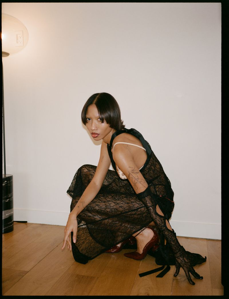 A slender adult female with straight dark hair and a sheer black dress crouching indoors on a wood floor against a white wall.