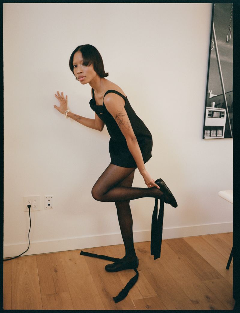 A slender woman in a black dress and pantyhose poses against a white wall while putting on a black shoe in an indoor setting.