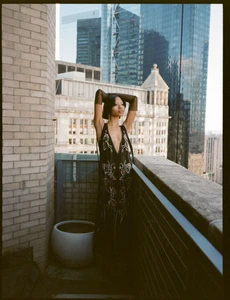 Model standing on a city rooftop with glass skyscrapers in the background, wearing a black embroidered gown with sheer sleeves.