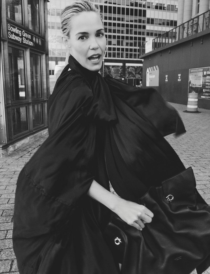 Adult woman in a black dress and holding a matching handbag stands in front of a subway station entrance and tall buildings in monochrome.