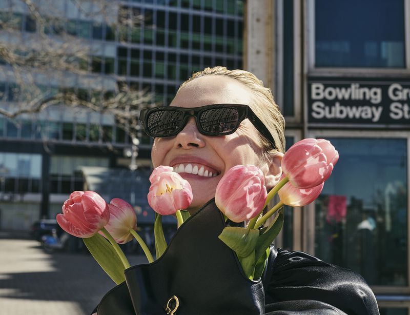 Woman smiles in the city sunlight, holding pink tulips in a black purse, wearing sunglasses with modern buildings and subway entrance.