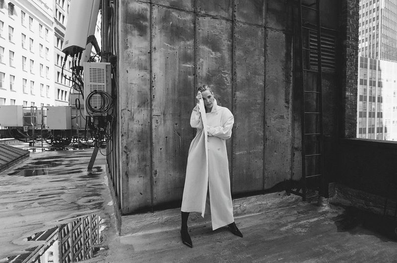 Woman in a white coat posing on a rooftop, black and white fashion photography in urban setting with city buildings in the background.